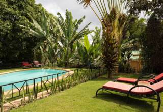 Secluded hotel pool surrounded by tropical plants with red cushioned loungers on a green lawn under cloudy sky