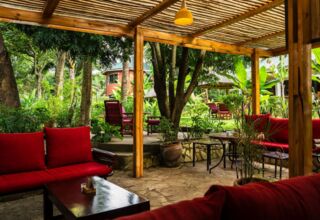 Outdoor lounge with red cushioned seating under a wooden pergola, surrounded by lush tropical garden and cozy seating areas