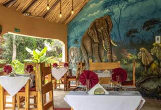 Open-air dining area with wooden furniture, white table linens, burgundy napkins, and a vibrant mural of elephants and lions