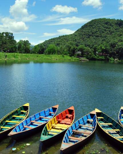 Colorful wooden boats lined up on a calm lake with lush green hills and a clear blue sky in the background