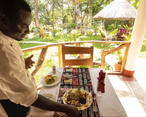Waiter serving a plated gourmet meal on a table with patterned runner on a garden terrace with thatched roof seating in tropical greenery