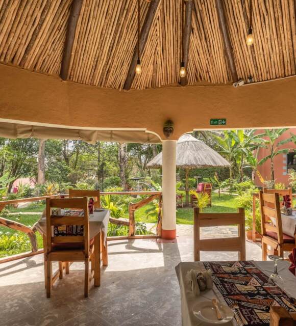 Open-air dining area with wooden chairs, patterned table runners, thatched roof, and lush tropical garden views