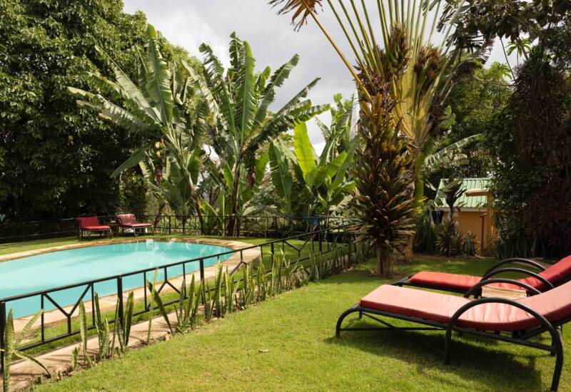 Secluded hotel pool surrounded by tropical plants with red cushioned loungers on a green lawn under cloudy sky