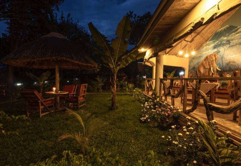 Outdoor dining area at night with thatched umbrellas, wooden chairs, candlelight, and a safari-themed wall mural featuring elephants