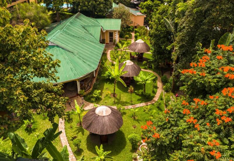 Lush tropical garden with winding paths, thatched umbrellas with seating, and a green-roofed hotel building surrounded by vibrant orange flowers.