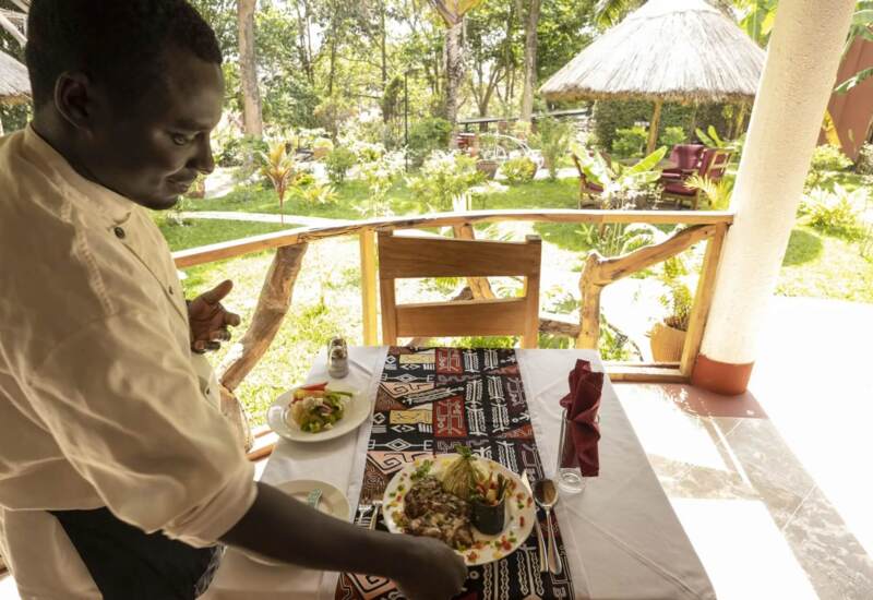 Server places a meal on a table with colorful patterned runner on a veranda overlooking lush garden with thatched roof seating areas