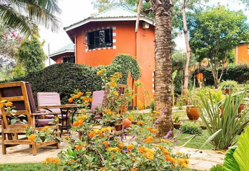 Outdoor seating with wooden chairs and cushions surrounded by vibrant flowers and tropical plants near orange garden cottages