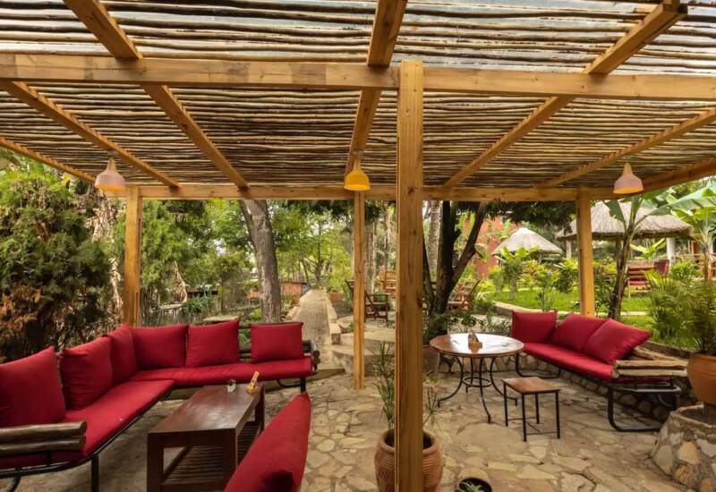 Outdoor seating area with red cushioned benches under a rustic wooden pergola, surrounded by lush greenery and stone flooring