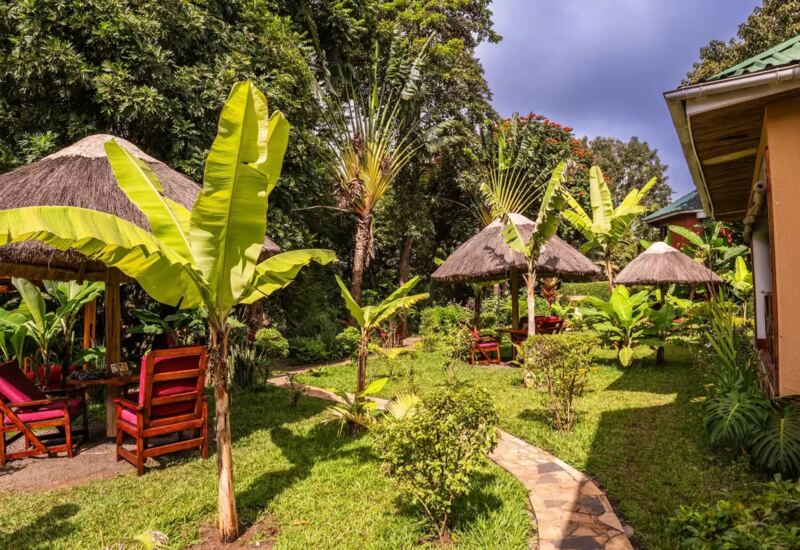 Tropical garden with thatched-roof seating huts, wooden chairs with red cushions, and lush greenery along stone pathways