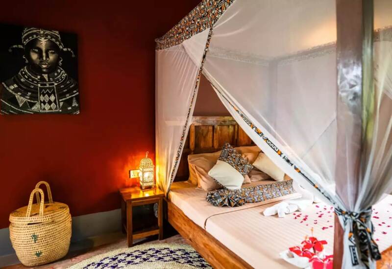 Cozy wooden four-poster bed with patterned mosquito netting and rose petals in a warm, stylish room with red walls and soft lighting.