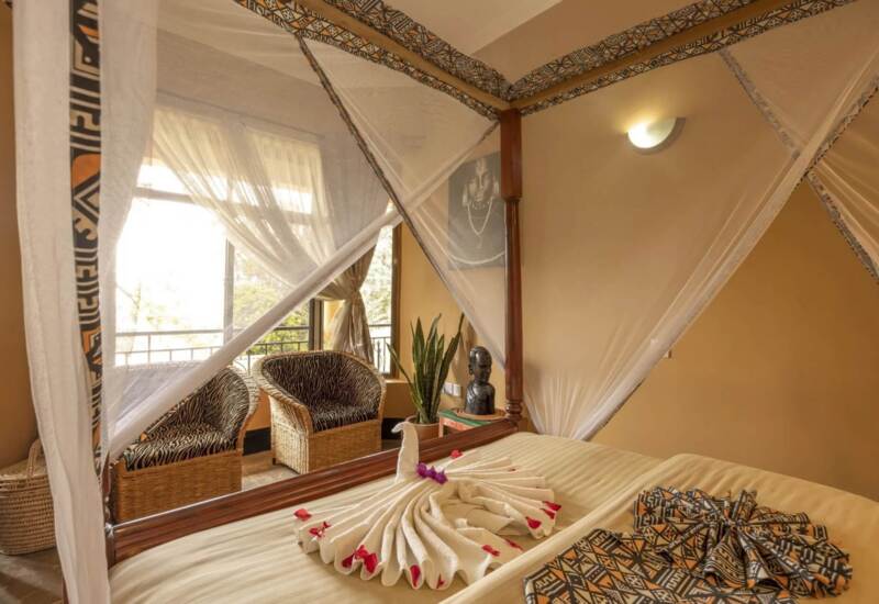Cozy hotel room with four-poster bed draped in mosquito net, patterned linens, wicker chairs, and natural light from balcony window