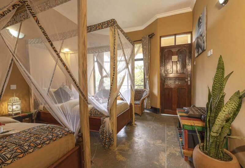 Cozy hotel room with four-poster bed draped in mosquito netting, patterned bedding, wicker chair, colorful bench, and large potted plant