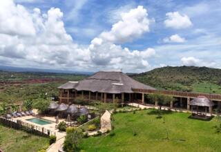 Thatched-roof lodge with outdoor pool, sun loungers, shaded cabanas, set in lush green hills under a partly cloudy blue sky