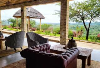 Open-air lounge with patterned armchairs and leather sofa overlooking lush greenery and distant hills under a thatched roof terrace