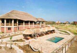 Thatched-roof lodge with an outdoor pool, sun loungers under straw umbrellas, and a hammock on a stone patio in a serene landscape.