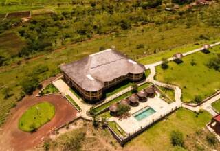 Thatched-roof lodge with outdoor pool and shaded seating, surrounded by green lawns and pathways in a savannah setting