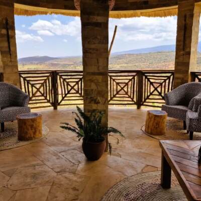Open-air lounge with patterned armchairs, wooden stools, and tribal decor overlooking expansive savanna and distant hills