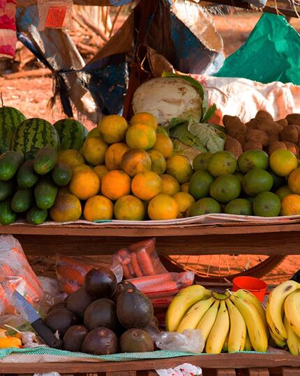 Outdoor rustic market stand displaying fresh bananas, oranges, watermelons, cucumbers, yams, and garlic under natural light