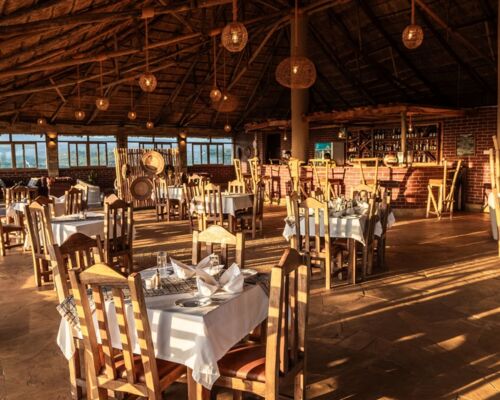 Rustic open-air restaurant with wooden chairs, white tablecloths, woven pendant lights, and a brick bar under a thatched roof