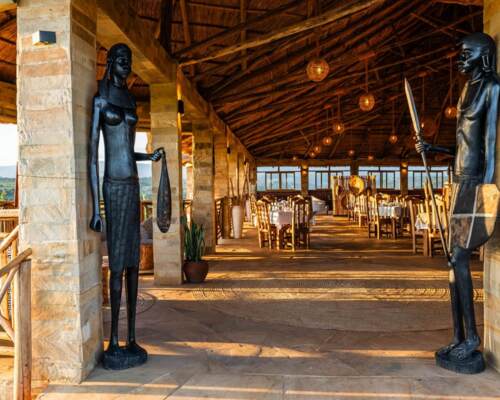 Open-air dining area with thatched roof, stone columns, wooden furnishings, and African-style statues at entrance, overlooking green landscape