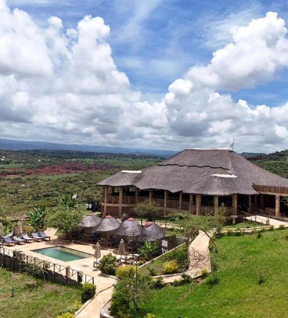 Thatched-roof lodge with outdoor pool, sun loungers, shaded cabanas, set in lush green hills under a partly cloudy blue sky