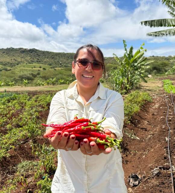 Smiling woman holding freshly picked red chili peppers in a lush farm garden with hills and blue sky background