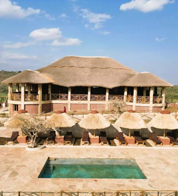 Thatched-roof lodge with rustic balcony overlooking a pool area with sun loungers and straw umbrellas, set in a scenic hillside location