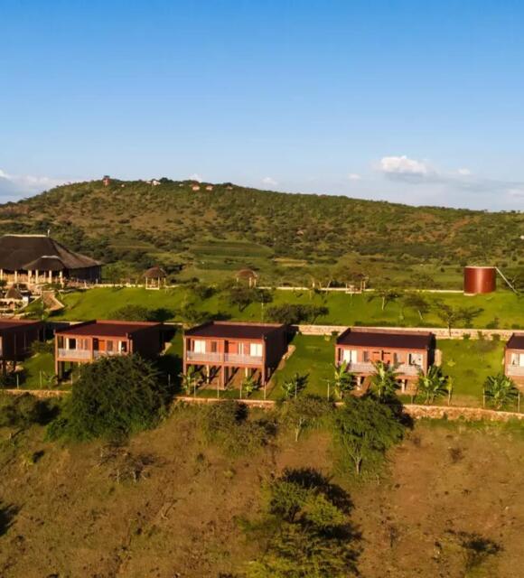 Row of elevated wooden lodges with balconies overlooking green hills and a large thatched-roof main building in a serene landscape
