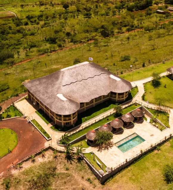 Thatched-roof lodge with outdoor pool and shaded seating, surrounded by green lawns and pathways in a savannah setting