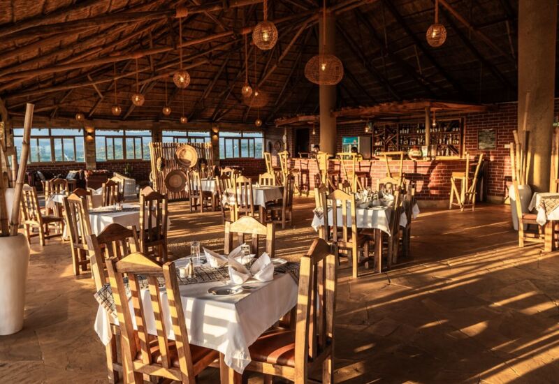 Rustic open-air restaurant with wooden chairs, white tablecloths, woven pendant lights, and a brick bar under a thatched roof