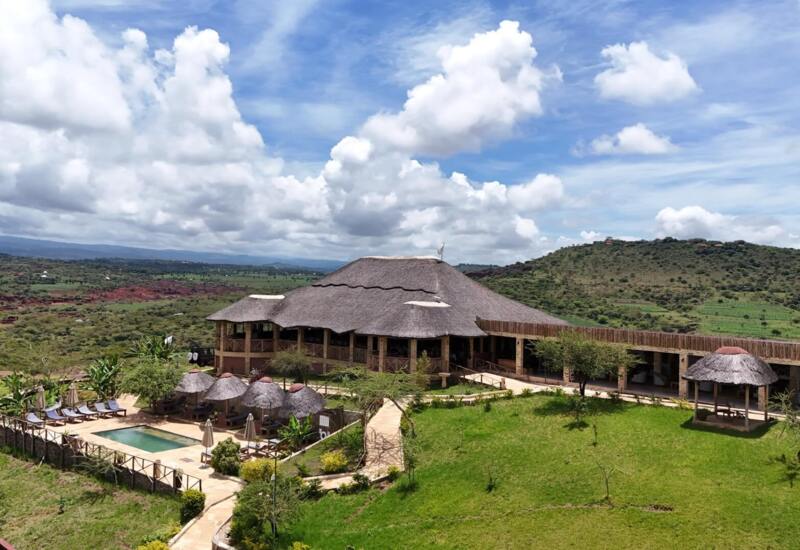 Thatched-roof lodge with outdoor pool, sun loungers, shaded cabanas, set in lush green hills under a partly cloudy blue sky