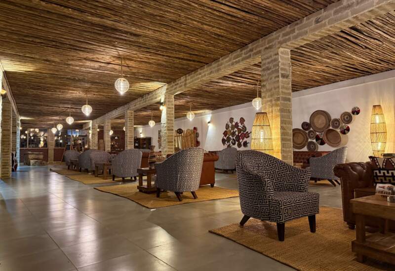 Spacious hotel lounge with patterned armchairs, warm wood ceiling, ambient lighting, woven wall decor, and cozy seating areas on rugs.