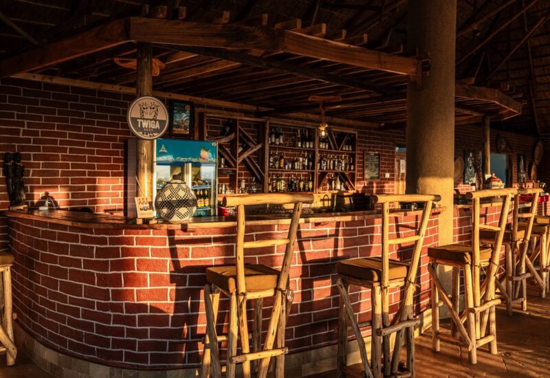 Rustic hotel bar with wooden stools, brick counter, thatched roof, and shelves stocked with liquor and drinks in warm sunlight