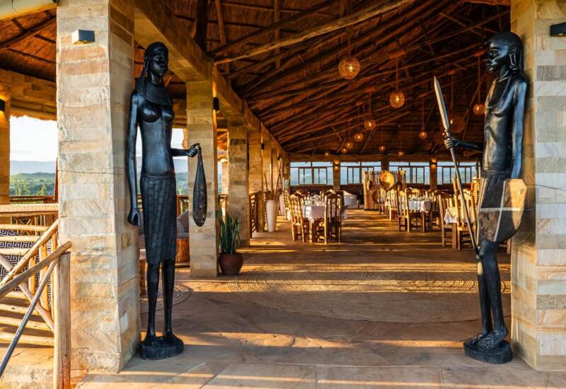 Open-air dining area with thatched roof, stone columns, wooden furnishings, and African-style statues at entrance, overlooking green landscape
