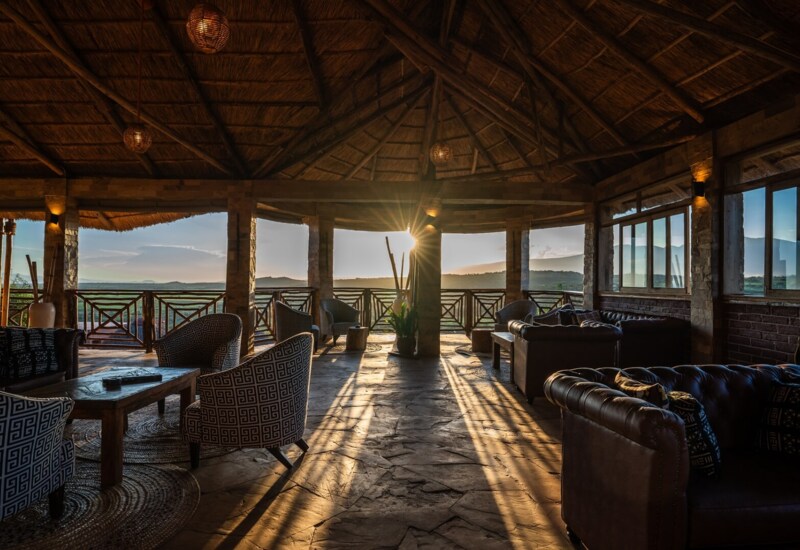 Rustic open-air lounge with thatched roof, cozy armchairs, leather sofas, and sunset views over rolling hills and valleys.