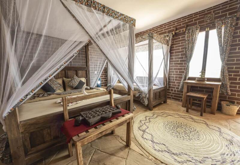 Rustic twin beds with mosquito netting, patterned curtains, and wooden furniture in cozy room with brick walls and woven rug.