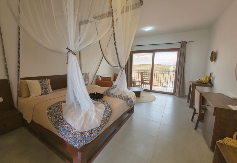 Spacious hotel room with a king bed draped in a decorative mosquito net, wood furnishings, balcony with scenic nature views, and cozy seating area