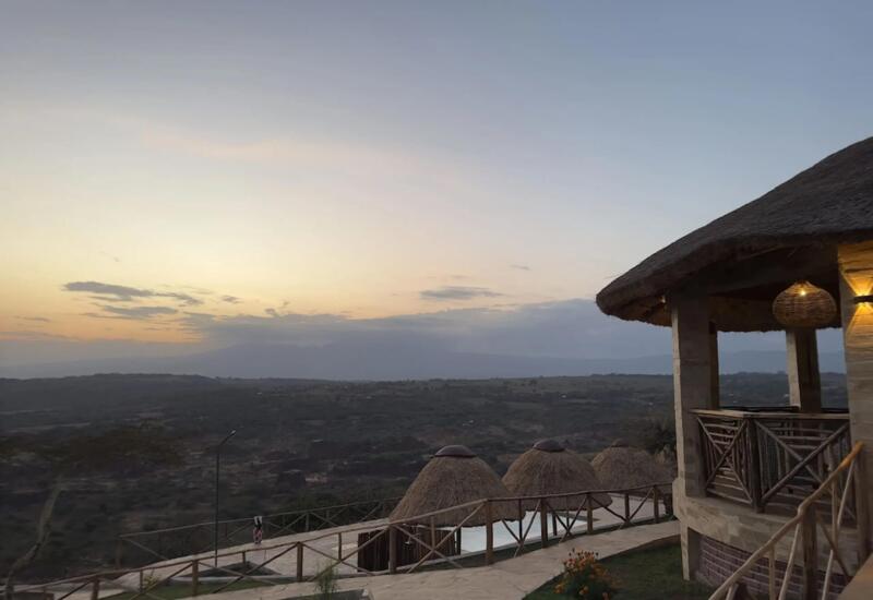 Thatched-roof lodge patio with warm lighting overlooking expansive hillside landscape at sunset, featuring rustic wooden railings