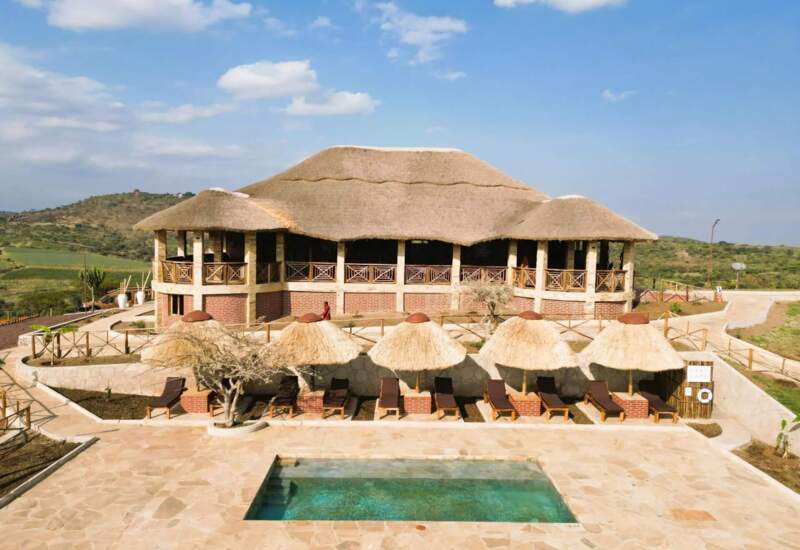 Thatched-roof lodge with rustic balcony overlooking a pool area with sun loungers and straw umbrellas, set in a scenic hillside location