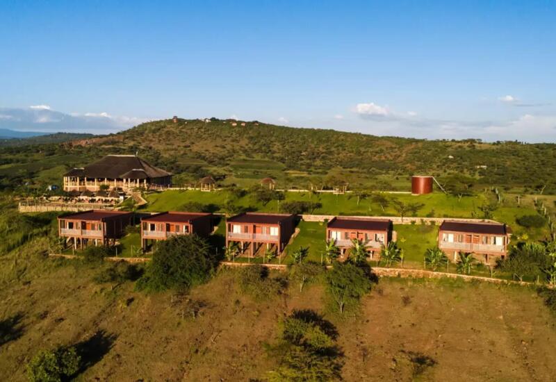 Row of elevated wooden lodges with balconies overlooking green hills and a large thatched-roof main building in a serene landscape