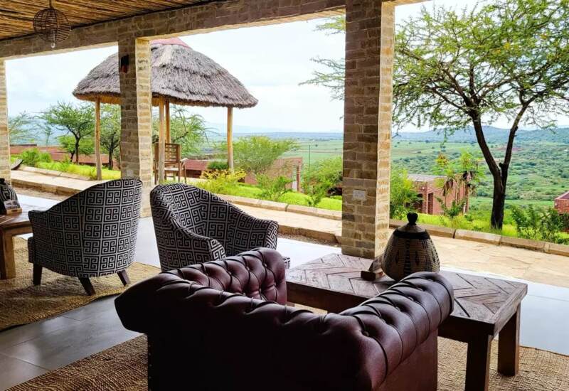Open-air lounge with patterned armchairs and leather sofa overlooking lush greenery and distant hills under a thatched roof terrace