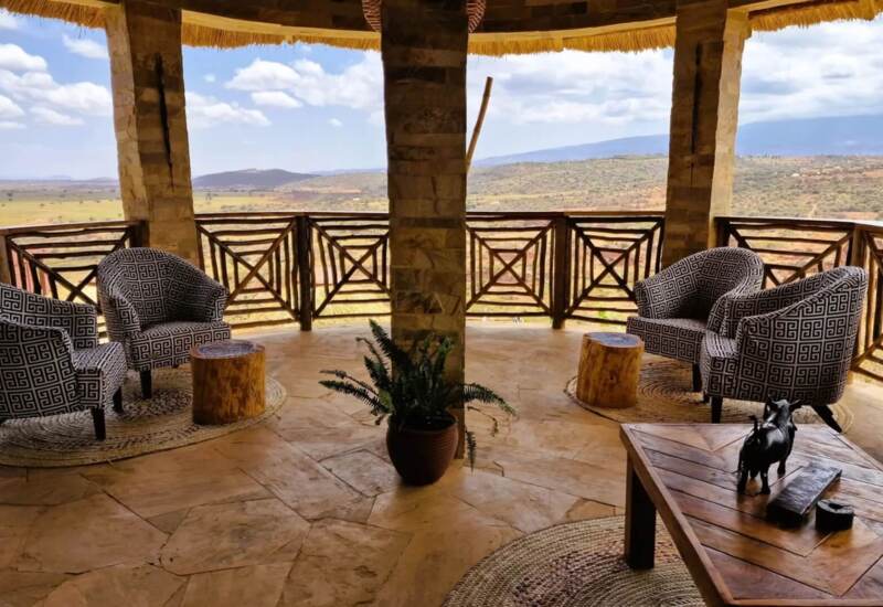 Open-air lounge with patterned armchairs, wooden stools, and tribal decor overlooking expansive savanna and distant hills