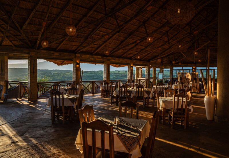 Open-air dining area with wooden tables, thatched roof, warm lighting, and scenic views of green hills at sunset