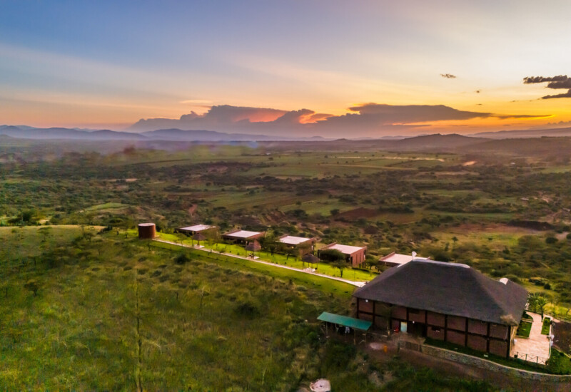 Secluded lodge with thatched roof and multiple cabins along a green path in vast savannah under a colorful sunset sky