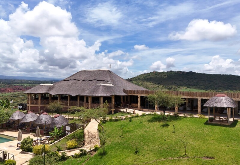 Thatched-roof lodge with outdoor pool, shaded loungers, and grassy gardens set against rolling green hills under a partly cloudy sky