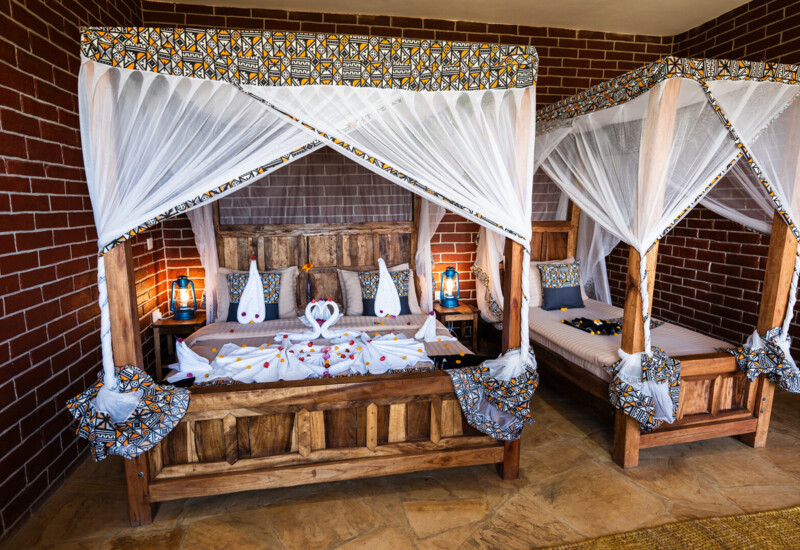Rustic twin wooden canopy beds with white mosquito nets and patterned trim in a cozy room with brick walls and lantern lighting
