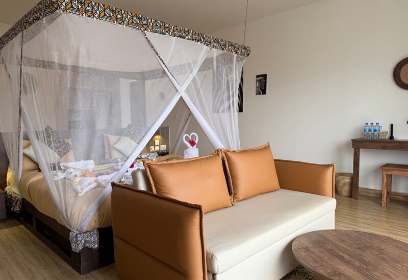 Cozy hotel room with a four-poster bed draped in decorative mosquito netting, leather sofa, wooden table, and simple furnishings