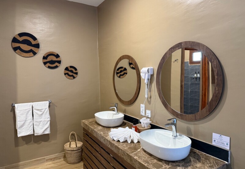 Hotel bathroom with dual white oval sinks, round wooden mirrors, beige walls, decorative woven wall art, and neatly folded towels