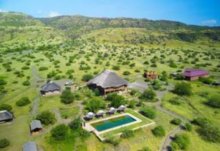 Eco-lodge with thatched roofs and a rectangular pool surrounded by lounge chairs, set in rolling green hills under a clear blue sky