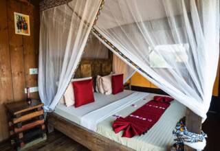 Cozy wooden room with a four-poster bed draped in white mosquito netting, accented by red and beige pillows and decorative runner.